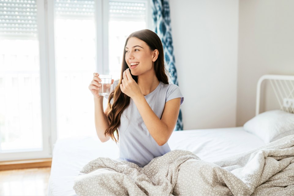 Person sitting on a bed holding a glass of water and a pill in a bright bedroom with patterned curtains.
