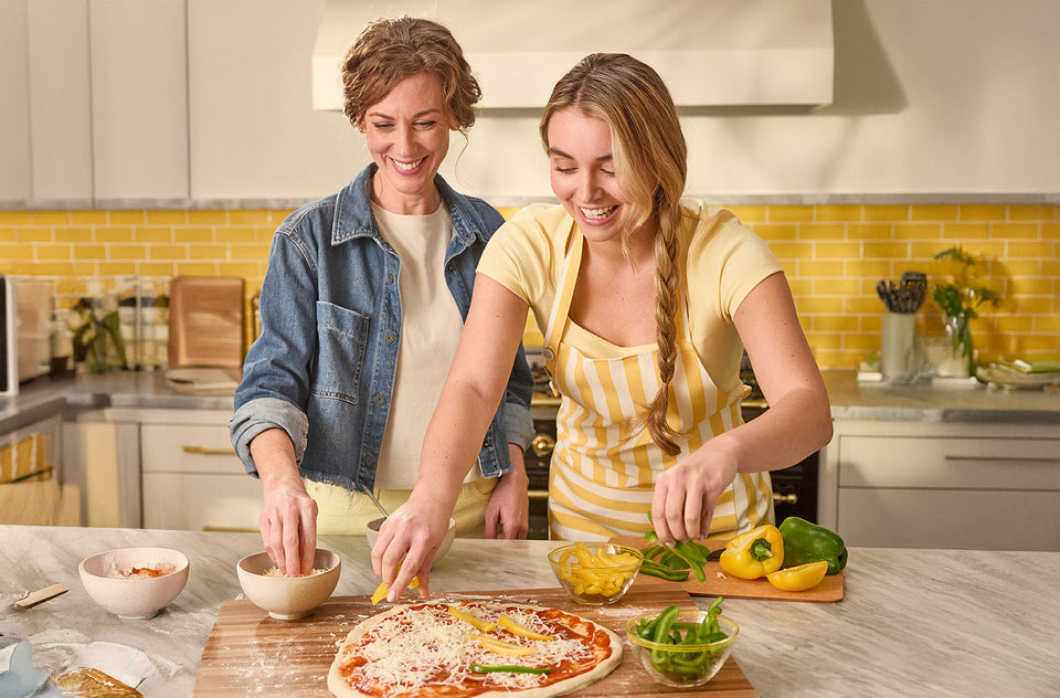 Two people in a kitchen adding toppings to a pizza on a wooden board.