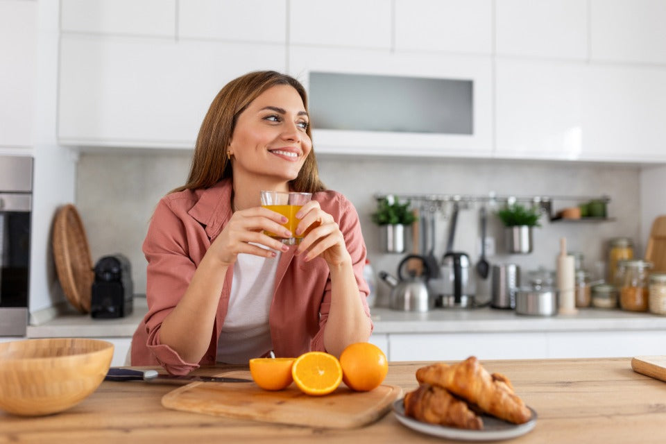 Person in a bright kitchen holding a glass of fresh orange juice with oranges and breakfast on the counter.