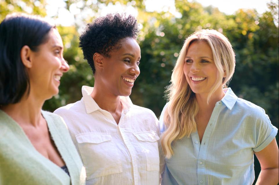 Three women smiling standing outdoors in light-colored shirts.
