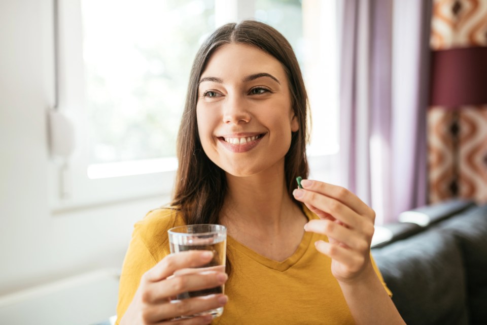 Woman smiling in a yellow shirt holding a glass of water in one hand and a supplement in the other, seated indoors near a window.