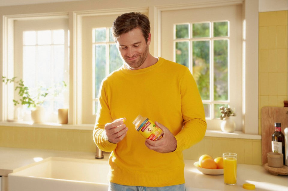 Person in yellow sweater holding a Nature Made Vitamin C gummies supplement bottle in a well lit kitchen.
