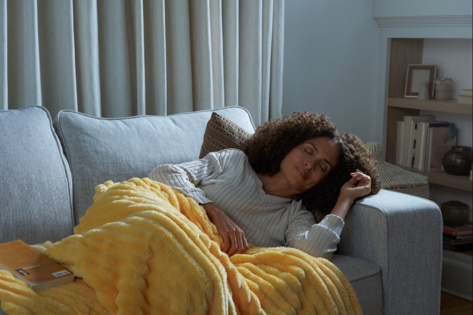 Person relaxing on a sofa under a cozy yellow blanket in a calm living room setting.