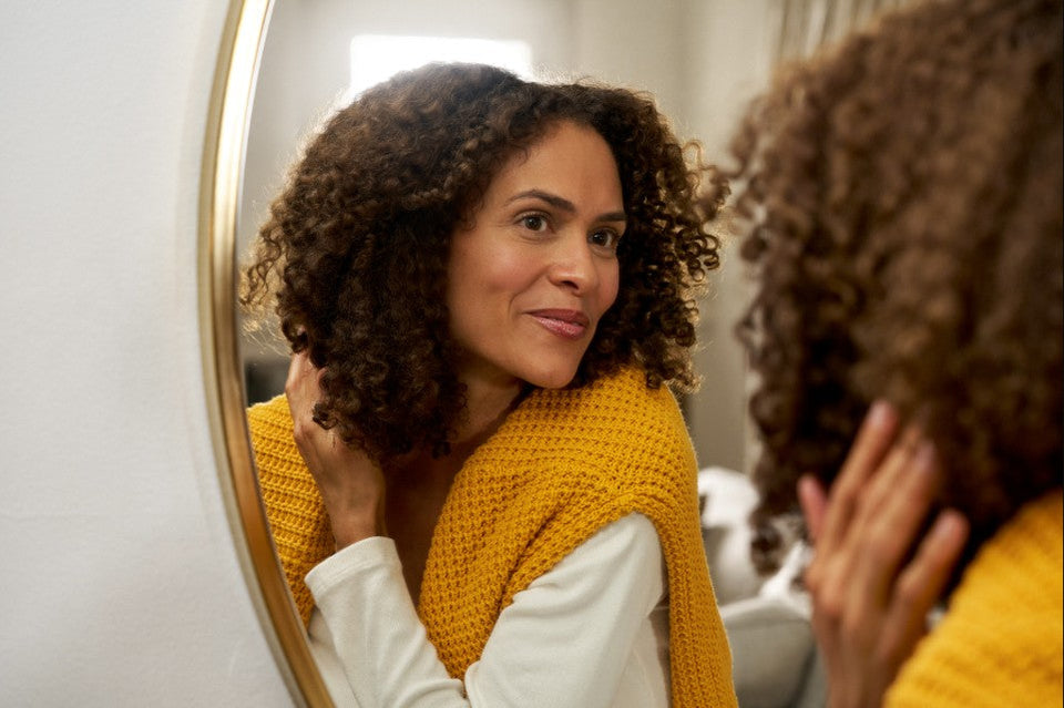 Person in yellow sweater touching their hair while looking in a mirror, illustrating hair care and nutrient support for hair health.