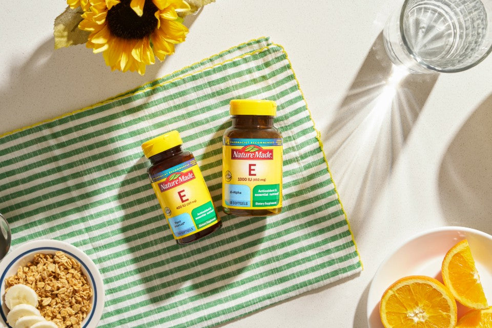 Nature Made Vitamin E bottles arranged on a table with fruit and water, representing daily vitamin E supplementation.
