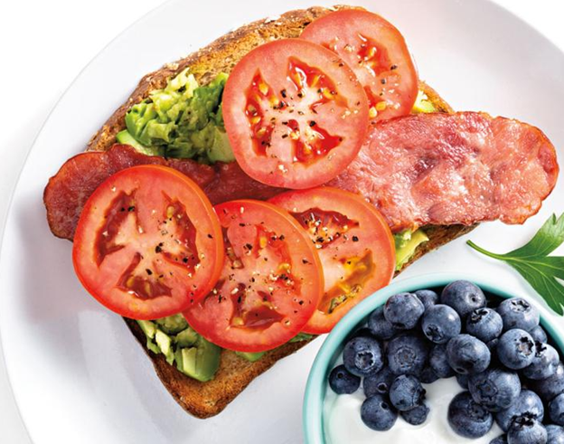 A plate of avocado toast with turkey bacon, tomato, and a side of blueberries and yogurt