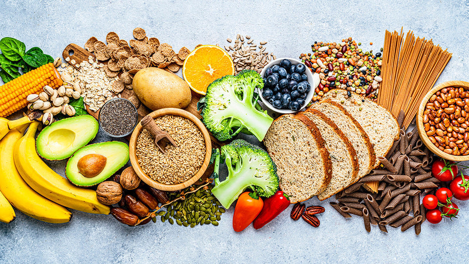 A colorful assortment of foods displayed on a light blue surface including Bananas, avocado, broccoli,  bread, berries, grains and nuts.