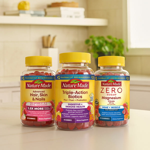 Three Nature Made gummy supplement bottles with yellow caps are lined up on a kitchen counter. The bottles include Advanced Hair, Skin & Nails; Triple-Action Biotics; and Zero Sugar Magnesium.