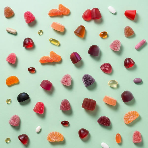 An overhead flat-lay arrangement of assorted gummy candies and softgel pills scattered across a light mint green background.
