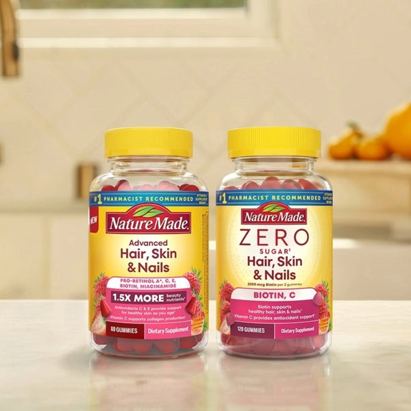 Two Nature Made supplement bottles on a countertop in a kitchen setting: Advanced Hair, Skin & Nails with a yellow cap and red gummies, and Zero Sugar Hair, Skin & Nails with a yellow cap and pink gummies.