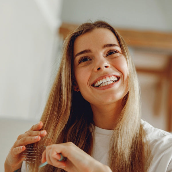 A smiling young woman with long light brown hair looks upward while brushing her hair with a dark brown paddle brush in a brightly lit indoor setting.