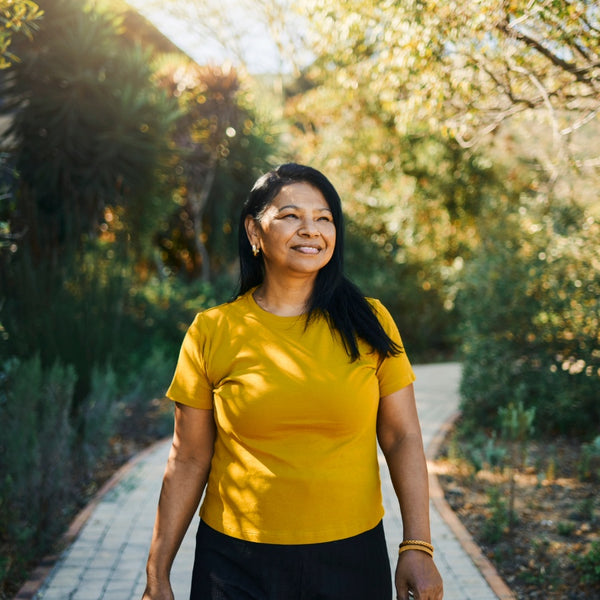 A woman with long black hair stands on a sunlit stone path wearing a bright yellow short-sleeve crewneck t-shirt and black pants, with a lush green garden background.