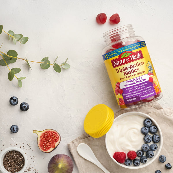 Nature Made Triple-Action Biotics dietary supplement bottle with a yellow lid, styled on a white textured surface with yogurt, blueberries, raspberries, figs, chia seeds, and eucalyptus leaves.