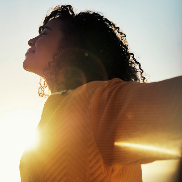 A woman with curly dark hair wearing a yellow top, backlit by a bright sun against a pale sky.