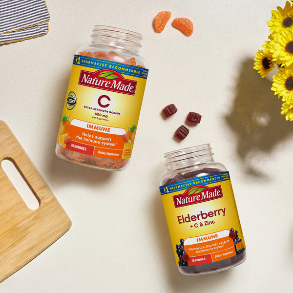 Two Nature Made C Immune and Elderberry + C & Zinc gummies supplement bottles on a light beige surface with  a wooden cutting board, yellow flowers, and loose gummies.