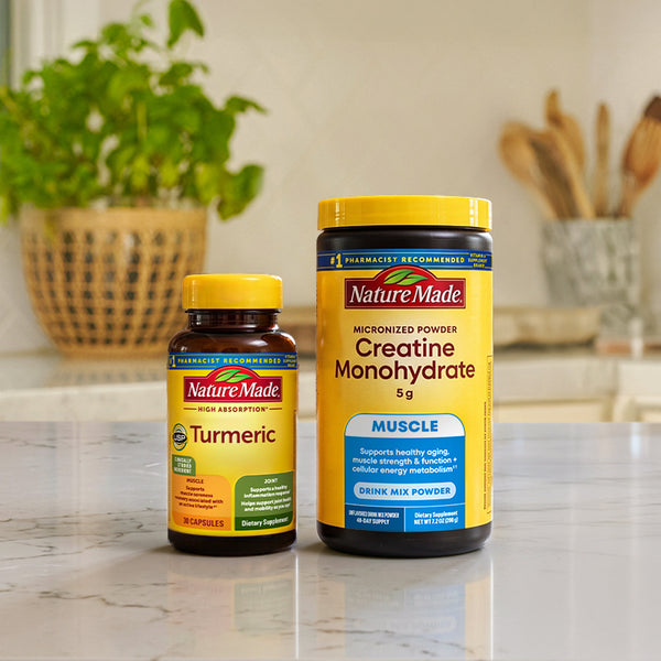 Two NatureMade dietary supplement containers on a marble countertop in a kitchen setting: a smaller Turmeric bottle and a larger Creatine Monohydrate powder tub, with blurred plants and kitchen items in the background.