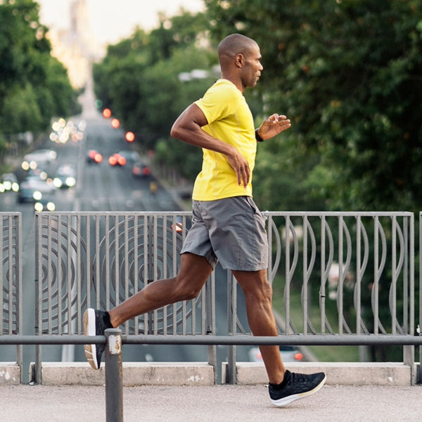 A man jogging on a city bridge wearing a bright yellow t-shirt, gray athletic shorts, and black running shoes, with blurred trees and street traffic in the background.