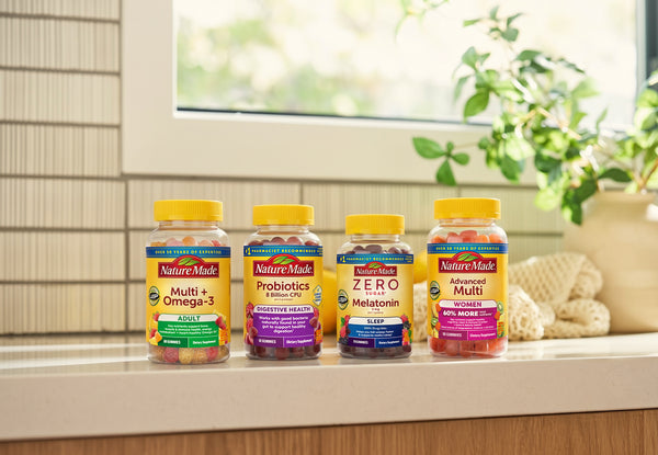 Four Nature-Made supplement bottles lined up on a kitchen counter in front of a window, with a potted plant and woven decor in the background. The bottles are Multi + Omega-3, Probiotics, Zero Melatonin, and Advanced Multi.