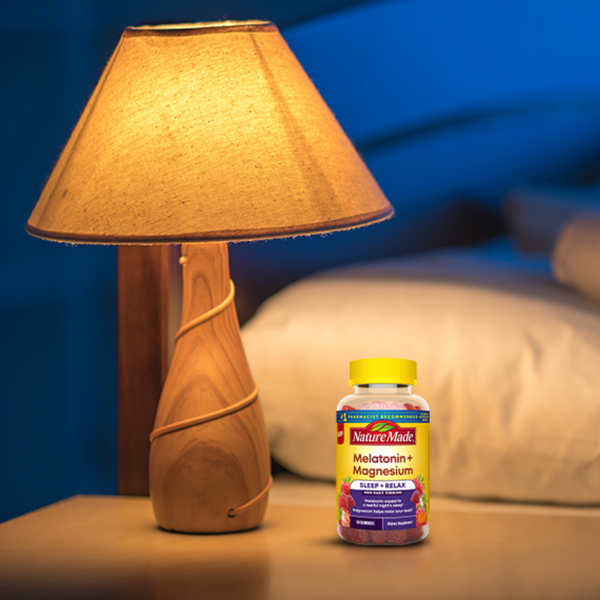 A bottle of Nature's Mask Melatonin + Magnesium sits on a bedside table next to a lit wooden table lamp in a cozy bedroom setting.