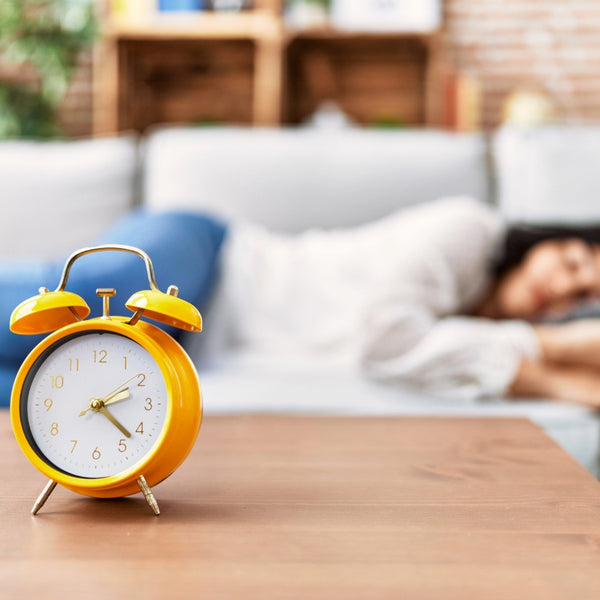 A bright yellow twin-bell alarm clock sits on a wooden table in the foreground, with a blurred living room scene behind it showing a person lying on a couch.