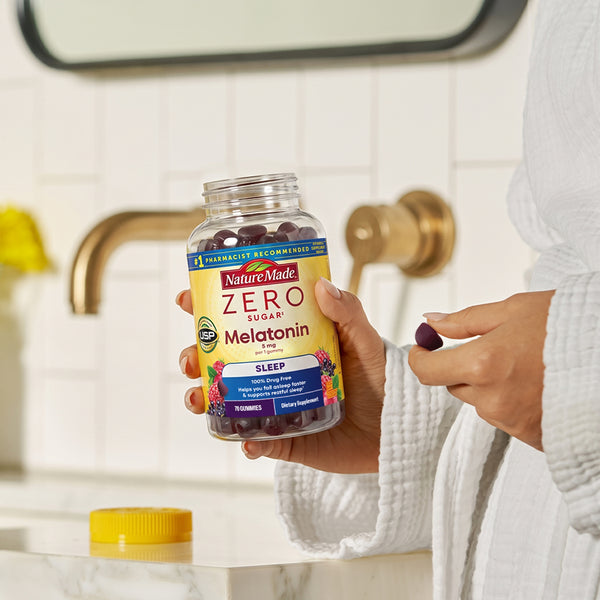 A person in a white robe holds a bottle of NatureMade Zero Sugar Melatonin Sleep gummies near a bathroom sink, with one hand holding a purple gummy and yellow top on the bathroom sink counter.
